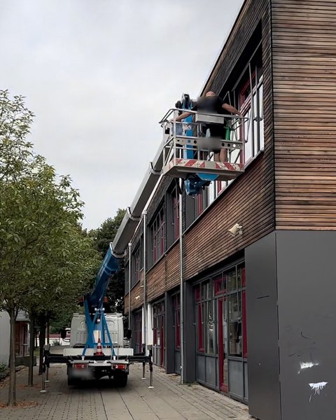 Zwei Mitarbeiter reinigen vom Arbeitskorb eines Hubsteigers die Fenster im Obergeschoss eines Gebäudes mit Holzfassade.