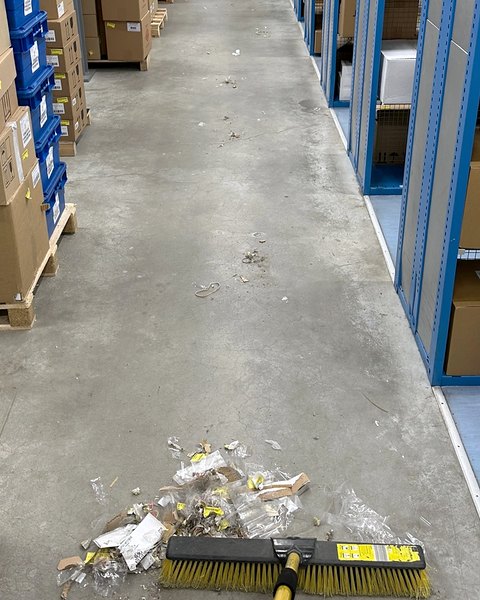 Lagergang zwischen Regalen, im Vordergrund kehrt ein Besen Folienreste und Kartonschnipsel zusammen.
