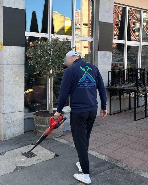 Ein Mitarbeiter im M&C-Pullover bläst mit einem Akku-Laubbläser den Gehweg vor einem Lokal sauber.
