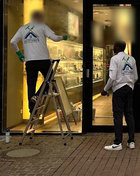 Zwei Mitarbeiter in M&C-Shirts reinigen am Abend die Schaufenster eines Geschäfts, einer davon auf einer Stehleiter.